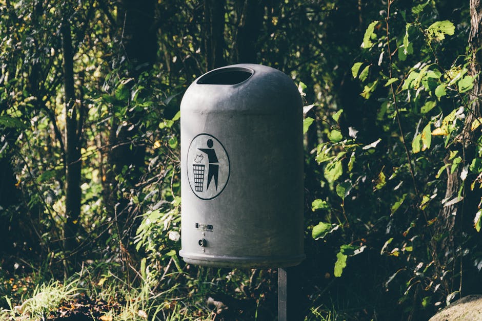 A cylindrical grey metal public waste bin with a rounded top and a large oval opening at the top for disposing of rubbish, positioned outdoors on a metal post. The front of the bin features a black pictogram icon showing a person throwing trash into a bin, and a small slot accessible for adding smaller waste items. The bin is situated amidst dense green foliage, including leaves and small branches, with natural sunlight filtering through the trees, creating a dappled light effect. The environmental setting appears to be a park or garden pathway, with the ground covered in grass and soil. The scene emphasizes the bin’s function for waste disposal in outdoor communal spaces, aligning with the context of independent rubbish collection and proper waste management carried out by waste collection services like Waste Collection Finchley, serving local residents and encouraging responsible disposal practices in natural environments.