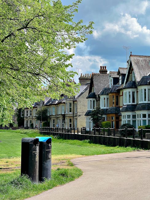 A row of traditional Victorian-style terraced houses with bay windows and slate roofs, situated along a suburban street on a bright day with partly cloudy skies. In the foreground, there are two black refuse bins with blue lids placed on the grassy verge beside a paved footpath. Behind the bins, a low black metal fence runs parallel to the houses, separating the individual front gardens from the footpath. A large, leafy deciduous tree with fresh green foliage extends its branches across the upper left corner of the image, casting partial shade on the grass and sidewalk. The environment appears well-maintained, with neatly kept lawns and tidy façades, indicative of a residential area that may utilize private refuse collection services, such as those provided by Waste Collection Finchley, for rubbish disposal. The overall scene emphasizes a quiet, well-organized community space that supports alternative waste handling methods outside of municipal collection routines.