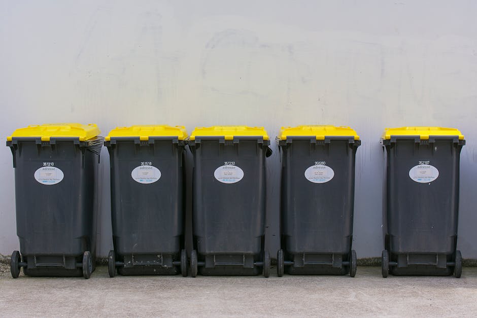 Five large wheeled rubbish bins made of dark grey plastic with textured surfaces are lined up on a concrete pavement against a plain, light-colored wall. Each bin has a yellow lid with a slightly ridged finish and a white label with black text and barcodes affixed to the front of the bins. The bins are positioned side by side, evenly spaced, and appear to be used for waste collection or recycling purposes. The background is a smooth, light surface with minimal details, and the lighting is even, highlighting the texture of the bins and the pavement. The scene suggests an outdoor area designated for waste disposal, possibly associated with private rubbish collection services such as Waste Collection Finchley, which may provide on-site clearance or alternative waste handling options in tight street environments.