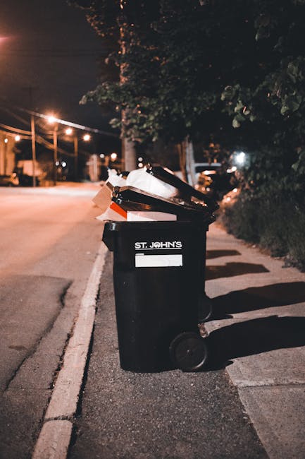 A black wheeled rubbish bin with a white label and the words 'ST JOHN'S' printed on the front, standing on a pavement next to a curb during evening hours. The bin is overflowing with assorted waste materials, including cardboard boxes and plastic bags, some of which are visibly leaning or protruding from the open lid. The surrounding environment shows a quiet street with dim street lighting, illuminated by warm glow from lampposts overhead. In the background, faint outlines of trees, parked cars, and residential buildings are visible, suggesting a suburban or urban area with limited space for waste collection. The scene reflects an instance of private or independent waste disposal, typical of a local rubbish removal service, such as Waste Collection Finchley, especially suitable for tight street conditions or on-site clearance where standard council refuse collection may be challenging. The overall atmosphere emphasizes a practical approach to rubbish management in residential areas, with a focus on straightforward waste disposal solutions suited to urban environments.