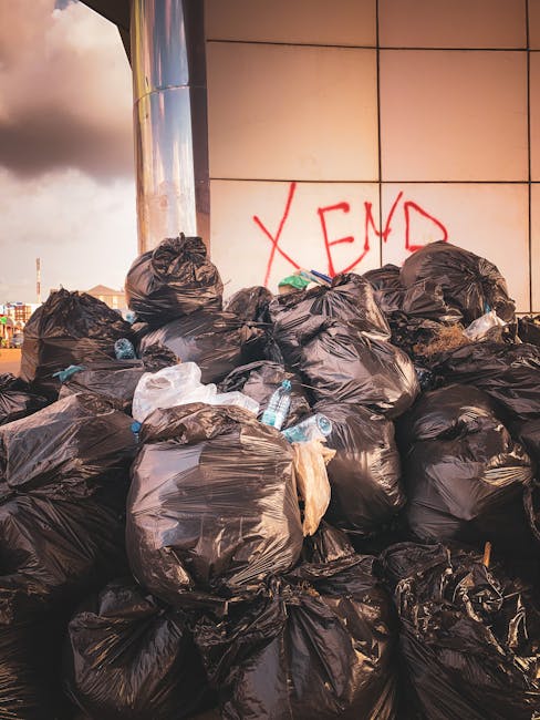 A large pile of black rubbish bags, filled and tied at the top, is stacked against a modern building with a tiled exterior wall. Some transparent plastic bottles and small waste items are visible protruding from the bags, indicating mixed household waste. The bags occupy the foreground and middle ground of the image, with the bags leaning and layered unevenly. Behind the pile, the building’s tiled wall features red graffiti that reads 'XEND' in spray paint. The scene appears to be outdoors, possibly on a pavement or driveway adjacent to the building. The lighting suggests late afternoon or early evening, with warm tones reflecting off the building's surface and the bags’ surfaces. The presence of waste suggests an instance of private waste accumulation needing collection, which Waste Collection Finchley might handle through their rubbish removal services, providing an alternative to municipal disposal methods. The urban environment and the nature of the waste imply an unsupported on-site clearance or individual disposal effort, rather than organized public collection.