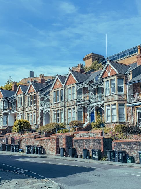 A row of Victorian-style terraced houses with ornate white-painted iron balcony railings and bay windows, situated along a sloped residential street. The houses are primarily constructed from red bricks with decorative detailing around the windows and gabled roofs with dark shingles. The front gardens are elevated behind low brick retaining walls and are landscaped with shrubs and small trees. In the foreground, a paved street features several black wheelie bins lined up along the curbside, indicating waste collection or disposal activity. The background includes modern buildings and a clear blue sky with some wispy clouds, suggesting a bright, sunny day. This street scene is indicative of an urban area where private waste management services, such as those offered by Waste Collection Finchley, might arrange alternative rubbish removal or on-site clearance beyond standard council waste collection. The overall environment is quiet and tidy, emphasizing clear access for rubbish removal and street maintenance.