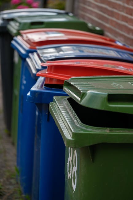 A large pile of black rubbish bags, filled and tied at the top, is stacked against a modern building with a tiled exterior wall. Some transparent plastic bottles and small waste items are visible protruding from the bags, indicating mixed household waste. The bags occupy the foreground and middle ground of the image, with the bags leaning and layered unevenly. Behind the pile, the building’s tiled wall features red graffiti that reads 'XEND' in spray paint. The scene appears to be outdoors, possibly on a pavement or driveway adjacent to the building. The lighting suggests late afternoon or early evening, with warm tones reflecting off the building's surface and the bags’ surfaces. The presence of waste suggests an instance of private waste accumulation needing collection, which Waste Collection Finchley might handle through their rubbish removal services, providing an alternative to municipal disposal methods. The urban environment and the nature of the waste imply an unsupported on-site clearance or individual disposal effort, rather than organized public collection.